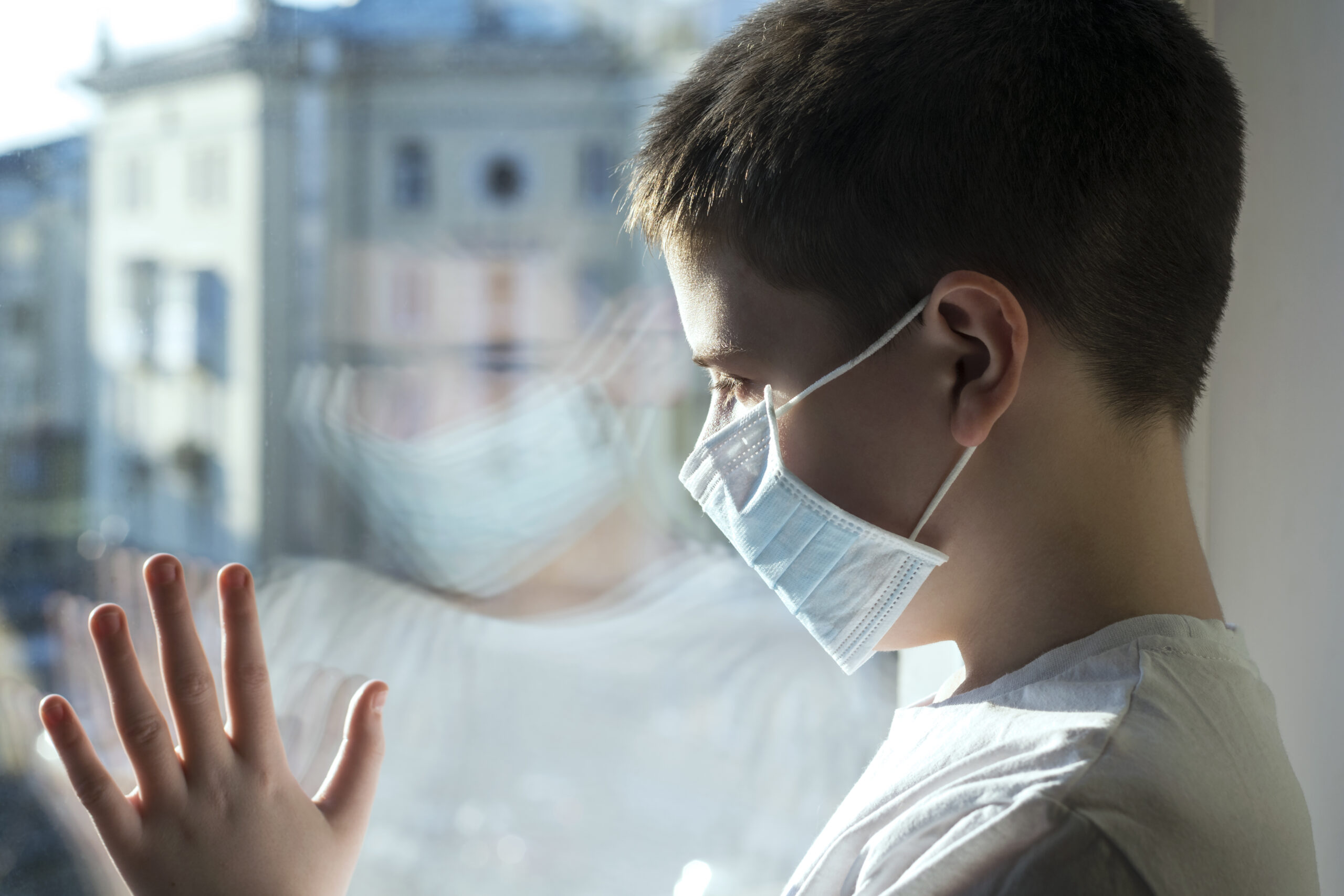 A boy in a medical mask looks wistfully out the window